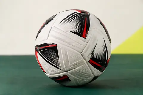 Ballon de football Mitre blanc, noir et rouge, posé sur un fond vert profond avec ses panneaux visibles
