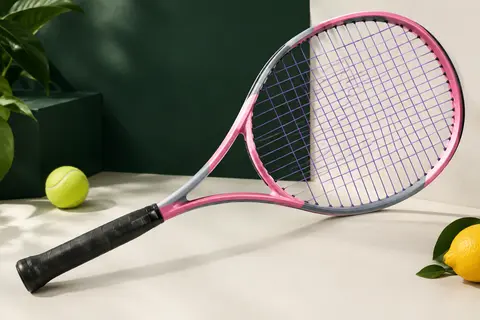 Raquette de tennis adulte rose et grise avec cordage violet, présentée sur un fond de studio vert profond.