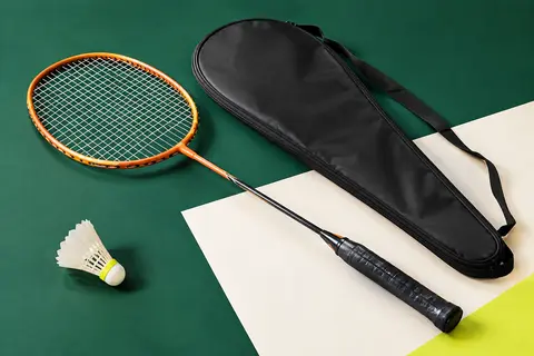 Raquette de badminton orange clair à tête isométrique, cadre aluminium et housse complète posée à côté.