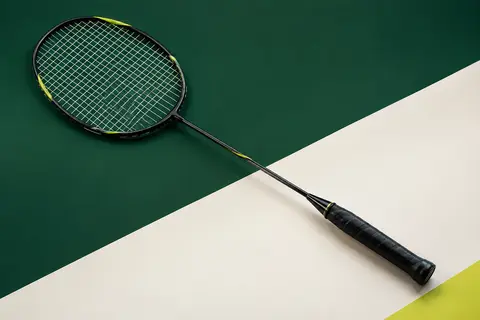 Raquette de badminton noire à tête isométrique, cordée, photographiée sur un fond de studio vert et blanc cassé.